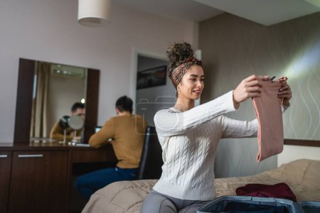 Foto de Woman caucasian couple packing or unpack wardrobe cloth from suitcase at hotel room while her husband or boyfriend sit in background happy smile travel and vacation concept copy space - Imagen libre de derechos