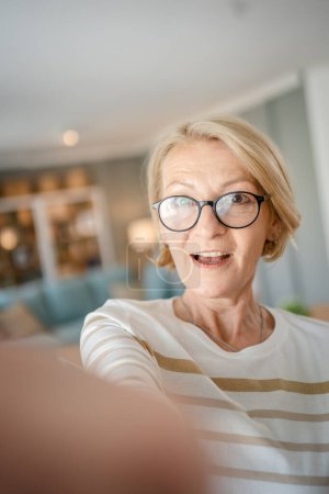 Téléchargez les photos : Portrait d'une femme caucasienne blonde mature avec des lunettes à la maison sourire heureux regardant vers la caméra pull de vêtements confiant dans la salle lumineuse utilisateur généré contenu autoportrait selfie ugc copie espace - en image libre de droit