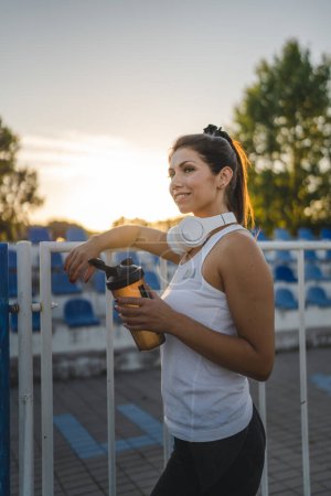 Téléchargez les photos : Une femme adulte caucasienne millénaire debout sur le stade sur la piste de course après ou avant l'entraînement en prenant un supplément de cale de frein agitateur bouteille d'eau style de vie sain concept copier l'espace - en image libre de droit