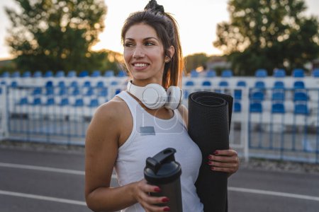 Foto de One woman adult caucasian female athlete standing on stadium on running track with supplement shaker and yoga mat with headphones ready for training outdoor in summer evening real people copy space - Imagen libre de derechos