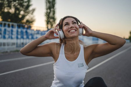 Photo for One happy caucasian woman brunette femaleathleete sitting on the running track in summer evening on stadium holding headphones listen music have fun while taking a brake real people copy space - Royalty Free Image