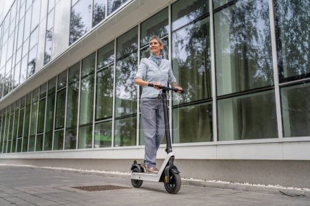 Photo for One woman mature senior caucasian female standing on electric kick scooter in day riding driving in town or city e-scooter eco friendly mode of transport real people copy space full length - Royalty Free Image
