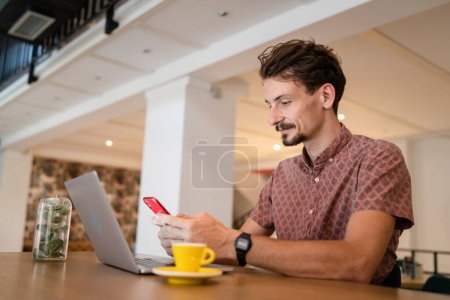 Foto de One caucasian man young adult millennial male using mobile phone sit at table in cafe at the table alone happy smile confident having online chat using internet for text messages while work remote - Imagen libre de derechos