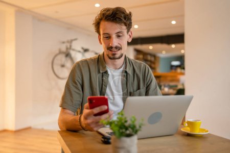Foto de One caucasian man young adult millennial male using mobile phone stand at table in cafe at the table alone happy smile confident having online chat using internet for text messages while work remote - Imagen libre de derechos