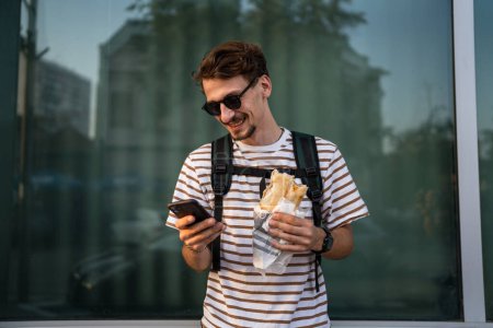 Téléchargez les photos : Un homme jeune adulte caucasien moderne mâle dans la ville dans la journée ensoleillée stand and eat sandwich fast food concept vie urbaine copier espace manger touristique en face de mur de construction personne réelle utiliser téléphone mobile - en image libre de droit