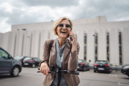 Photo for One woman mature caucasian blonde female stand outdoor with electric kick push scooter use mobile phone smartphone to make a call talk in the city stand alone confident real people copy space - Royalty Free Image