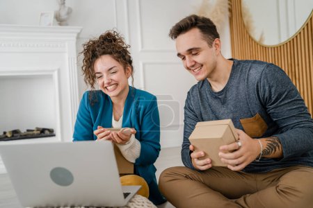 Photo for Young caucasian couple woman and man wife and husband or girlfriend and boyfriend opening gifts box presents and read card in front of laptop computer at home having online video call happy copy space - Royalty Free Image