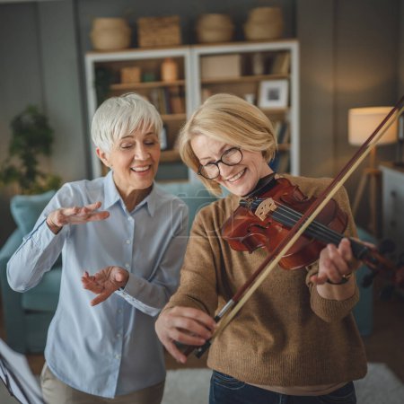 Photo for Two women mature female teacher give private violin lesson to her student or friend at home help her to learn to play in room hobby and leisure active senior concept copy space - Royalty Free Image