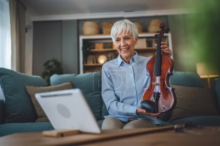 Photo for Mature senior caucasian woman learn to play violin practice at home online professor teacher teach or have internet private class on digital tablet while sit on sofa bed at home active senior hobby - Royalty Free Image
