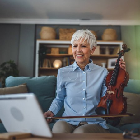 Photo for Mature senior caucasian woman learn to play violin practice at home online professor teacher teach or have internet private class on digital tablet while sit on sofa bed at home active senior hobby - Royalty Free Image