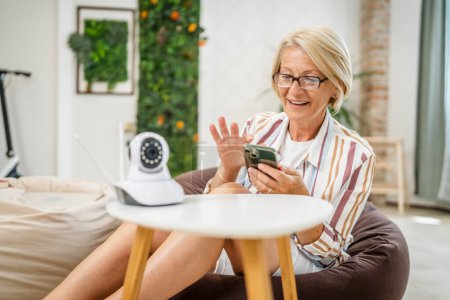 Photo for One mature caucasian woman holding home modern security surveillance camera and mobile phone trying to install and adjust connect an app copy space - Royalty Free Image