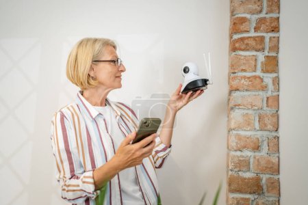 Téléchargez les photos : Une femme caucasienne mature tenant à la maison une caméra de surveillance de sécurité moderne et un téléphone mobile essayant d'installer et d'ajuster connecter un espace de copie d'application - en image libre de droit