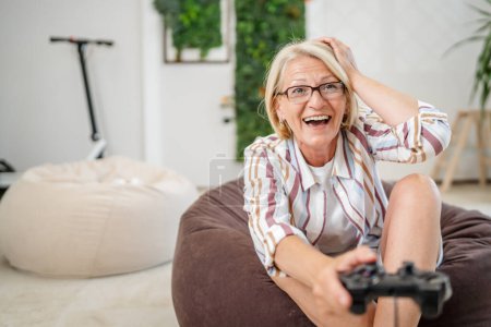 Téléchargez les photos : Une femme blonde caucasienne mature avec des lunettes jouer console de jeu vidéo en utilisant un contrôleur joystick tout en étant assis à la maison personnes réelles loisirs concept copier espace heureux sourire célébrer gagner de l'espace de copie - en image libre de droit