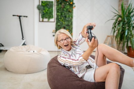 Foto de Una mujer rubia caucásica madura con anteojos jugar consola de videojuegos usando controlador de joystick mientras está sentado en casa personas reales concepto de ocio copiar espacio sonrisa feliz celebrar ganar espacio de copia - Imagen libre de derechos