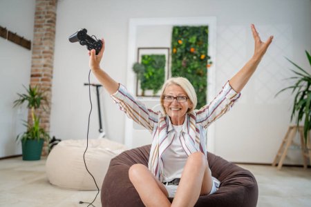 Photo for One mature caucasian blonde woman with eyeglasses play video game console using joystick controller while sitting at home real people leisure concept copy space happy smile celebrate win copy space - Royalty Free Image
