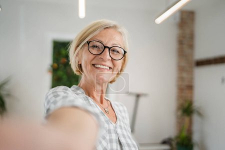 Téléchargez les photos : Portrait d'une femme caucasienne blonde mature avec des lunettes à la maison sourire heureux regardant vers la caméra pull de vêtements confiant dans la salle lumineuse utilisateur généré contenu autoportrait selfie ugc copie espace - en image libre de droit