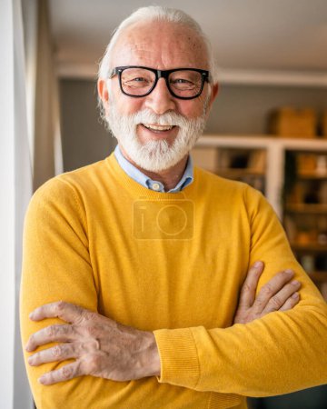 Téléchargez les photos : Portrait d'un homme âgé avec barbe et cheveux gris porter des lunettes heureux sourire positive émotion copie espace debout à la maison intérieur pull jaune - en image libre de droit