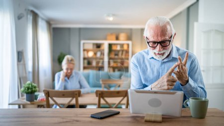 Téléchargez les photos : Homme âgé caucasien vieux grand-père mâle assis à la maison utiliser une tablette numérique pour l'application d'appel vidéo en ligne parler tandis que la femme s'asseoir derrière des personnes réelles hors foyer espace de copie concept de loisirs famille - en image libre de droit