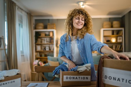 Photo for One young adult woman caucasian female with curly hair prepare wardrobe clothes for charity donation help at home putting stuff in the boxes and checking them out - Royalty Free Image