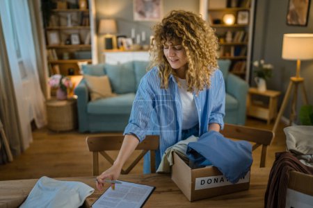 Photo for One young adult woman caucasian female with curly hair prepare wardrobe clothes for charity donation help at home putting stuff in the boxes and checking them out - Royalty Free Image