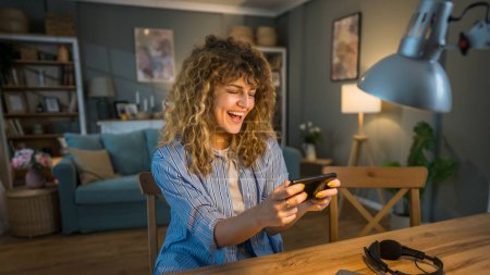 Photo for One adult caucasian woman with curly hair sit at home happy smile play video games while take a brake from work leisure activity having fun hold mobile phone use smartphone have fun online copy space - Royalty Free Image