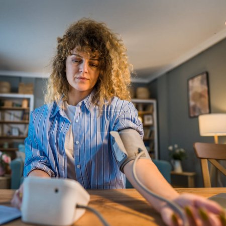 Téléchargez les photos : Une femme caucasienne pigiste assise à la table pendant le contrôle de travail mesurer la pression artérielle à la maison concept de soins de santé personne réelle - en image libre de droit