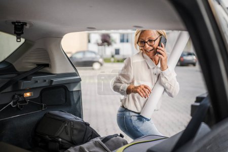 Photo for One mature blonde beautiful business woman travel concept female talk on smartphone take stuff belongings from the back of her car while moving or arrive to destination or work real people copy space - Royalty Free Image