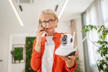 Photo for One mature caucasian woman holding home modern security surveillance camera and mobile phone trying to install and adjust connect an app copy space - Royalty Free Image