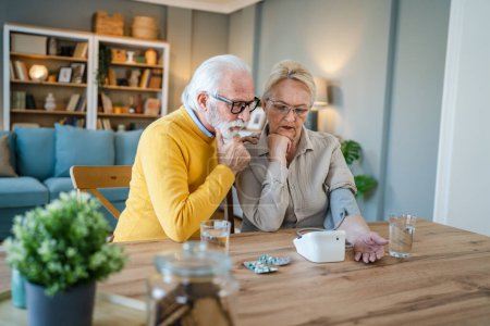 Téléchargez les photos : Couple âgé femme vérifier mesurer la pression artérielle tandis que son mari vieil homme caucasien assis à côté de la table à la maison de vraies personnes famille personnes âgées retraite et concept de santé - en image libre de droit