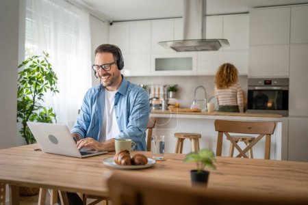 Photo for One man caucasian male freelancer work from home with headphones on his head on laptop computer customer support happy smile confident his wife stand in the background - Royalty Free Image