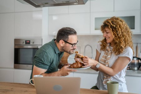 Téléchargez les photos : Caucasien couple homme et femme prendre le petit déjeuner dans la cuisine manger croissant et café tous les jours matin routine domestique vie de famille personnes réelles copier l'espace - en image libre de droit