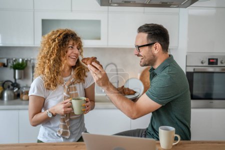 Photo for Caucasian couple man and woman having breakfast in the kitchen eat croissant and coffee daily morning routine domestic family life real people copy space - Royalty Free Image