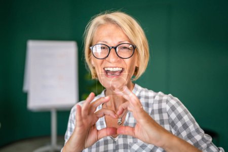 Téléchargez les photos : Portrait d'une femme caucasienne blonde mature avec des lunettes à la maison ou au bureau sourire heureux regardant vers l'espace de copie confiant appareil photo - en image libre de droit