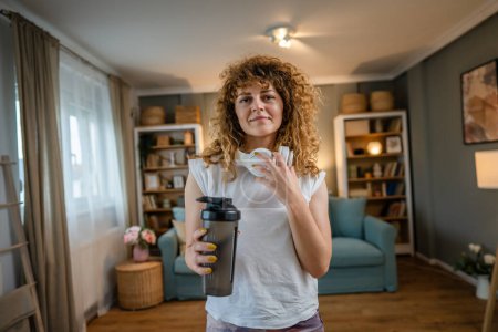 Téléchargez les photos : Portrait de femme adulte caucasienne avec des cheveux bouclés stand à la maison tenir shaker supplément porter t-shirt blanc avant ou après l'entraînement mode de vie sain concept copier l'espace - en image libre de droit