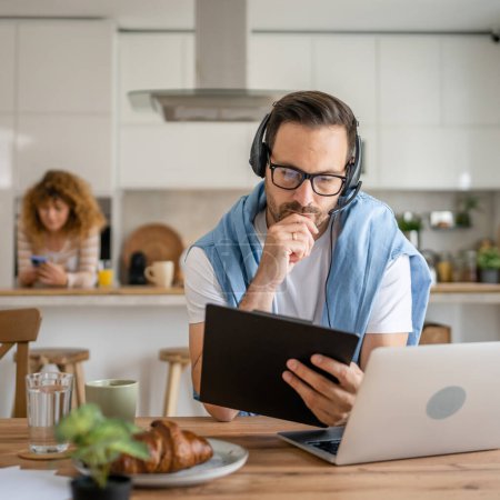 Photo for One man caucasian male freelancer work from home with headphones on his head on laptop computer customer support thinking serious his wife stand in the background - Royalty Free Image