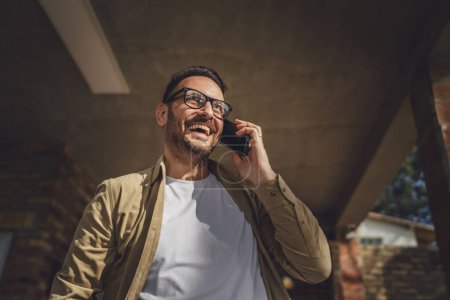 Téléchargez les photos : Un homme adulte caucasien mâle avec barbe et lunettes de vue en plein air dans la journée ensoleillée sourire heureux utiliser téléphone intelligent mobile pour faire un appel parler copie espace vraies personnes - en image libre de droit