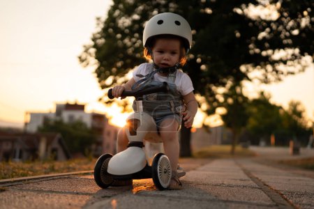 Téléchargez les photos : Small caucasian girl toddler playing outdoor in summer evening in sunset with 3 wheel children's kick scooter kid wear protective helmet real people copy space leisure family growing up concept - en image libre de droit