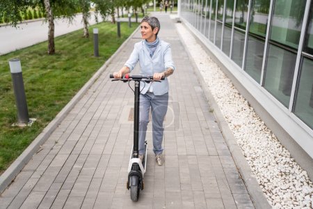 Téléchargez les photos : Une femme mature femme caucasienne âgée debout sur scooter électrique coup de pied dans la conduite d'équitation de jour en ville ou en ville e-scooter mode de transport respectueux de l'environnement personnes réelles copier l'espace pleine longueur - en image libre de droit