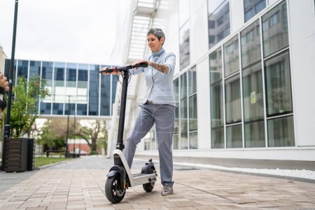 Photo for One woman mature senior caucasian female standing outdoor with electric kick scooter in day confident businesswoman using alternative mode of transport real people copy space - Royalty Free Image