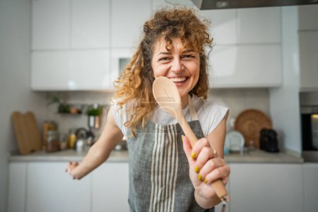Foto de Una joven mujer caucásica adulta feliz usar delantal en la cocina sonrisa mantener mezcla cuchara cantar - Imagen libre de derechos