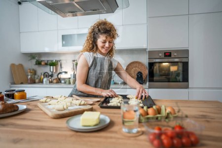 Téléchargez les photos : Une jeune femme caucasienne adulte heureuse porter tablier dans la cuisine sourire préparer pâte croissant snacks petit déjeuner - en image libre de droit
