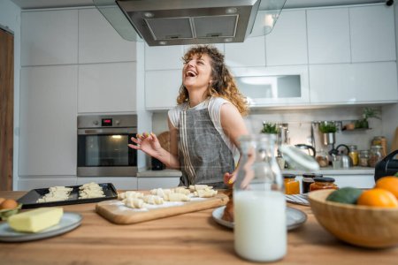 Foto de Una joven mujer caucásica adulta feliz usar delantal en la sonrisa de la cocina preparar masa croissant snacks desayuno - Imagen libre de derechos
