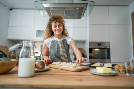 Photo for One happy young adult caucasian woman wear apron in the kitchen smile prepare dough croissant breakfast snacks - Royalty Free Image