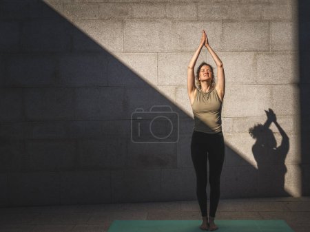 Téléchargez les photos : Une femme caucasienne adulte pratiquant le yoga en plein air jour d'été dans la ville en face du mur avec ombre et soleil mode de vie sain concept de bien-être auto-soin - en image libre de droit