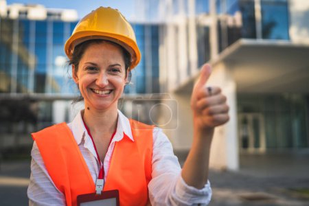 Foto de Arquitecto mujer mujer ingeniero de construcción de pie al aire libre usar casco protector y al oeste en frente de la pared del edificio moderno - Imagen libre de derechos