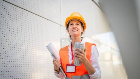 Foto de Arquitecto mujer mujer ingeniero de construcción de pie al aire libre usar casco protector y al oeste en frente de la pared del edificio moderno - Imagen libre de derechos
