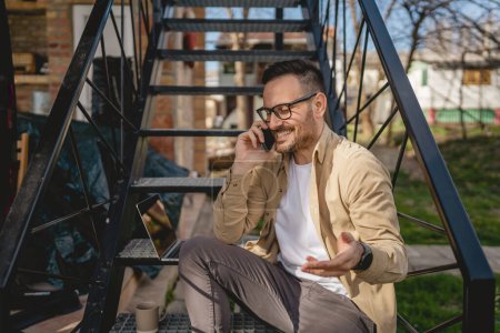 Photo for One man caucasian male sit on the steps outdoor while taking a brake from work with laptop use mobile phone to make a call talking happy smile confident real person - Royalty Free Image
