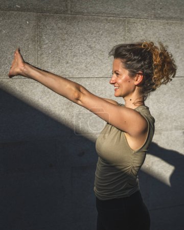 Téléchargez les photos : Une femme caucasienne adulte faisant du yoga pratique étirer en plein air jour d'été dans la ville en face du mur avec ombre et soleil mode de vie sain concept de bien-être auto-soins - en image libre de droit