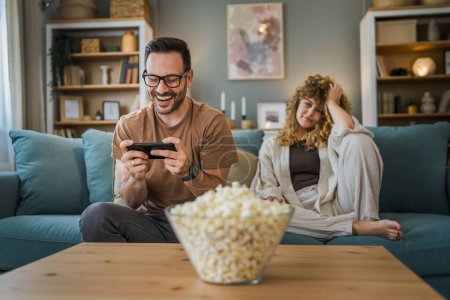 Téléchargez les photos : Homme adulte caucasien mâle assis sur le canapé-lit à la maison jouer à des jeux vidéo sur smartphone tenir téléphone mobile tandis que sa femme ou sa petite amie en arrière-plan est déçu - en image libre de droit
