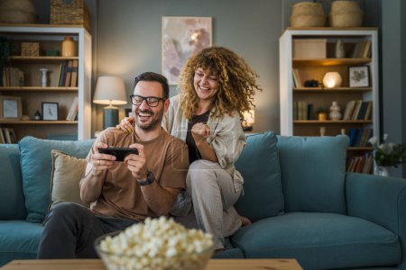 Photo for Adult couple man and woman caucasian husband and wife or boyfriend and girlfriend he play video games at home hold mobile phone smart phone have fun leisure joy and bonding concept copy space - Royalty Free Image